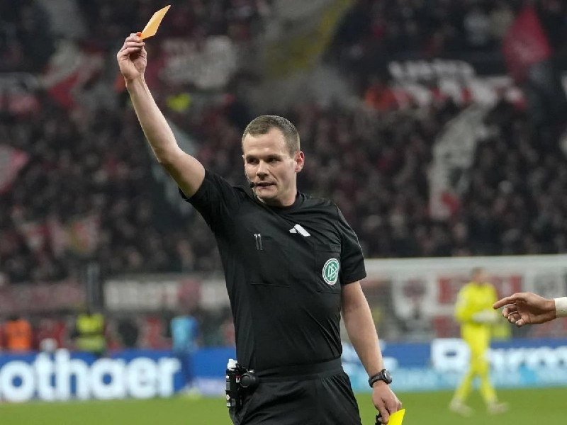 A referee in a black uniform holds up a yellow card in his right hand on a soccer field, with blurred spectators in the background.