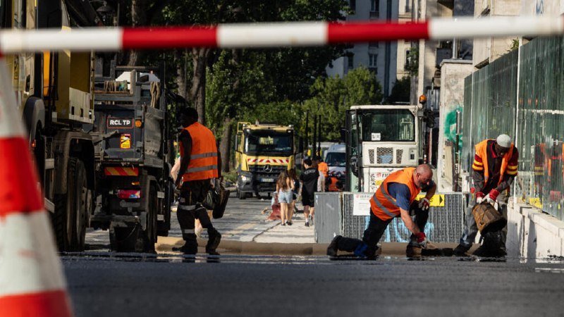 Workers in orange high-visibility vests perform road maintenance on a street, with heavy machinery and a red-and-white barrier visible in the foreground.