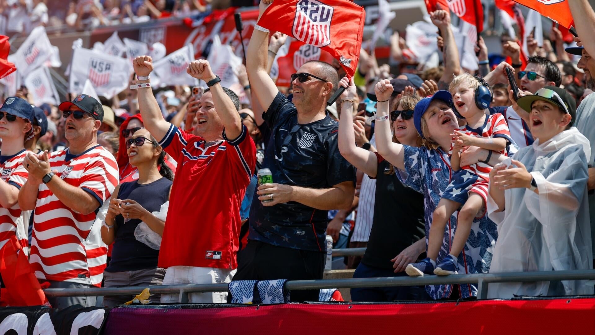 USA soccer fans in red, white, and blue gear cheer and wave USA flags from stadium stands, with a young child wearing noise-canceling headphones.
