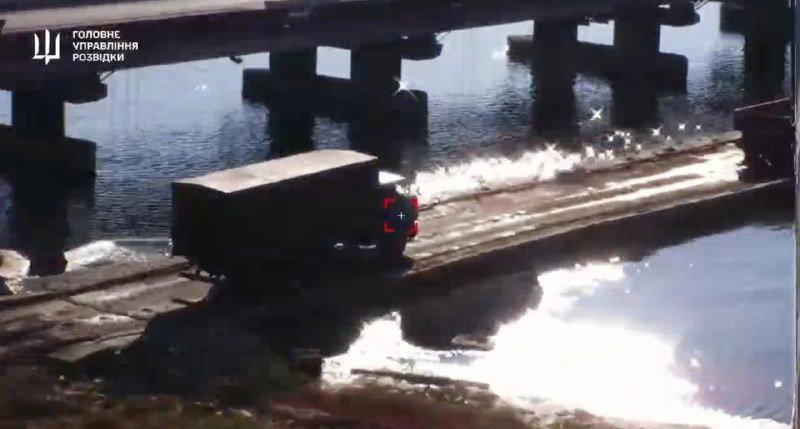 A military truck crosses a water-spanning bridge, with a Ukrainian intelligence logo and Cyrillic text visible in the upper left corner.
