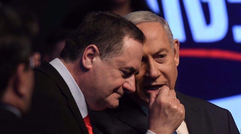 Two men in dark suits speak closely together at a political event, with partial Hebrew lettering visible on a screen behind them.