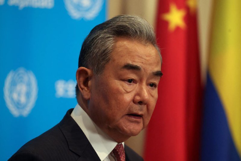 A gray-haired man in a dark suit and red tie speaks at a podium, with a blue UN emblem backdrop and Chinese and other flags visible behind him.