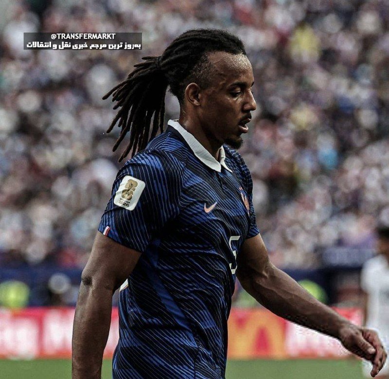 A soccer player with dreadlocks wearing a dark blue striped jersey with a white collar stands on a pitch before a blurred stadium crowd.