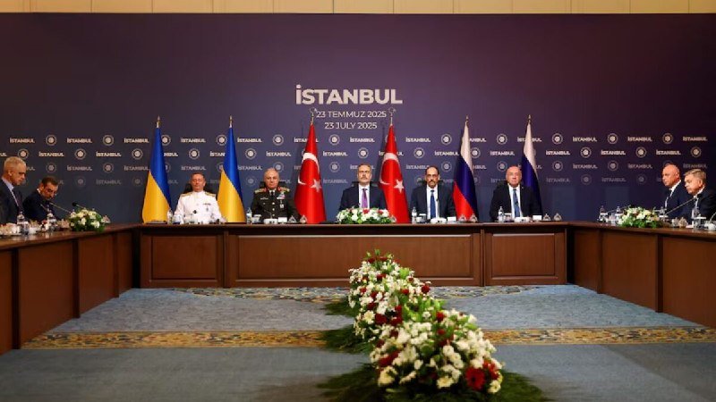 Several officials in suits and military uniforms sit at a long conference table adorned with flowers, with Ukrainian, Turkish, and Russian flags displayed against a backdrop reading "İSTANBUL."