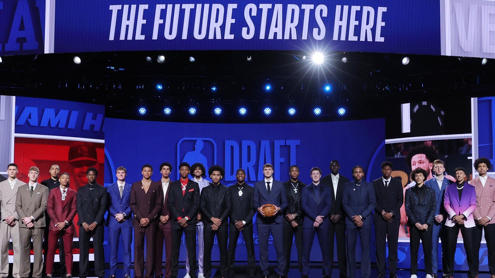 A group of young men in suits stand on a stage beneath a banner reading "THE FUTURE STARTS HERE," with an NBA Draft logo displayed behind them.