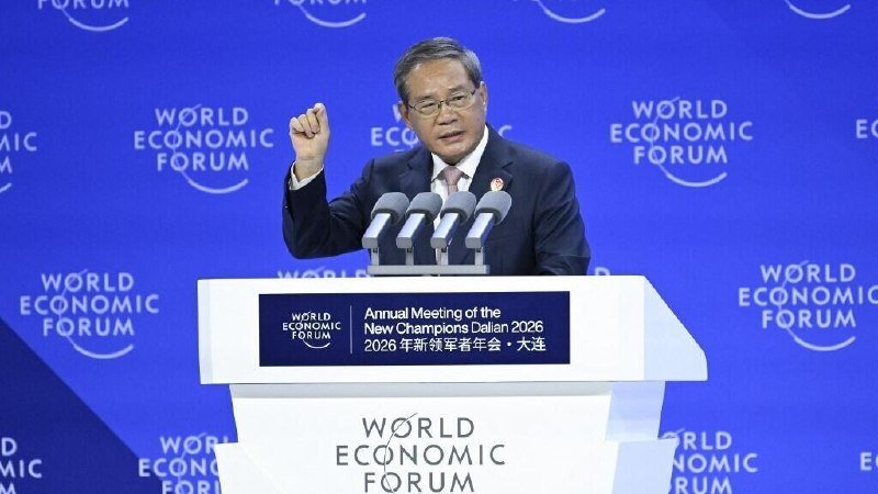 A man in a dark suit and glasses speaks at a podium with a raised fist, set against a blue backdrop reading "World Economic Forum" and "Annual Meeting of the New Champions Dalian 2026."