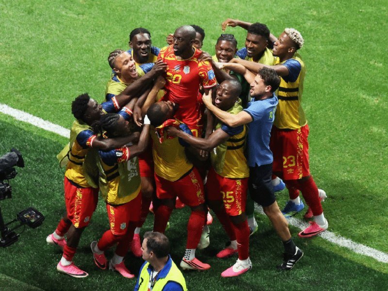 Soccer players in yellow and red jerseys celebrate together in a jubilant group huddle on a green field.