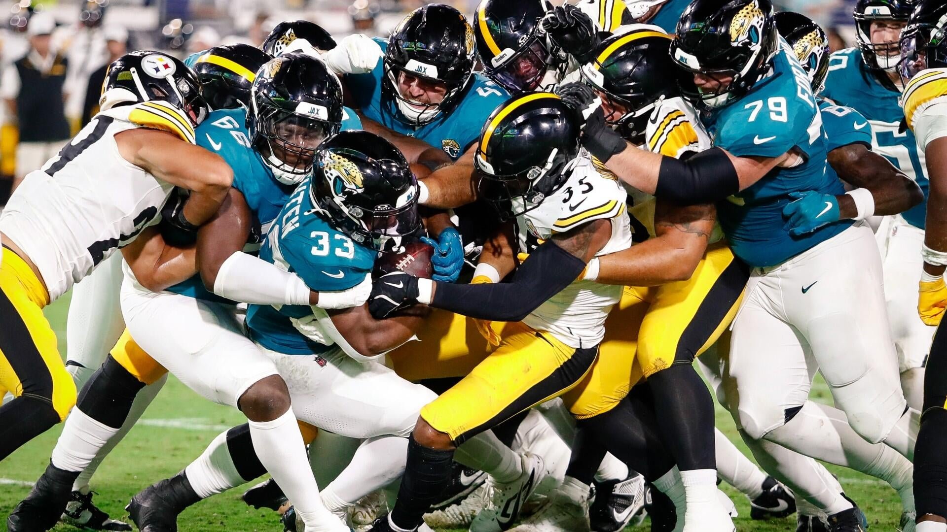 Football players in teal and white uniforms clash with players in black and yellow uniforms as they pile up during a tackle on a grass field.