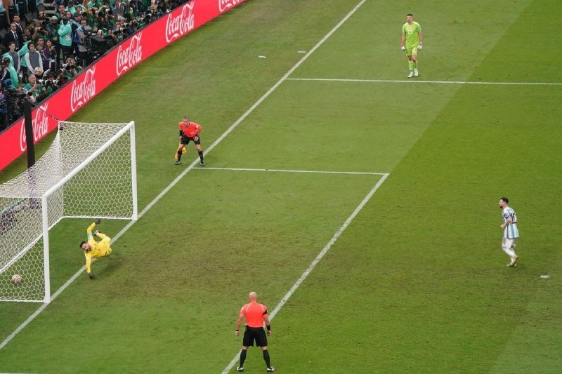 A goalkeeper in yellow dives as a soccer ball crosses the goal line, with players and a referee watching during a match featuring Coca-Cola signage.
