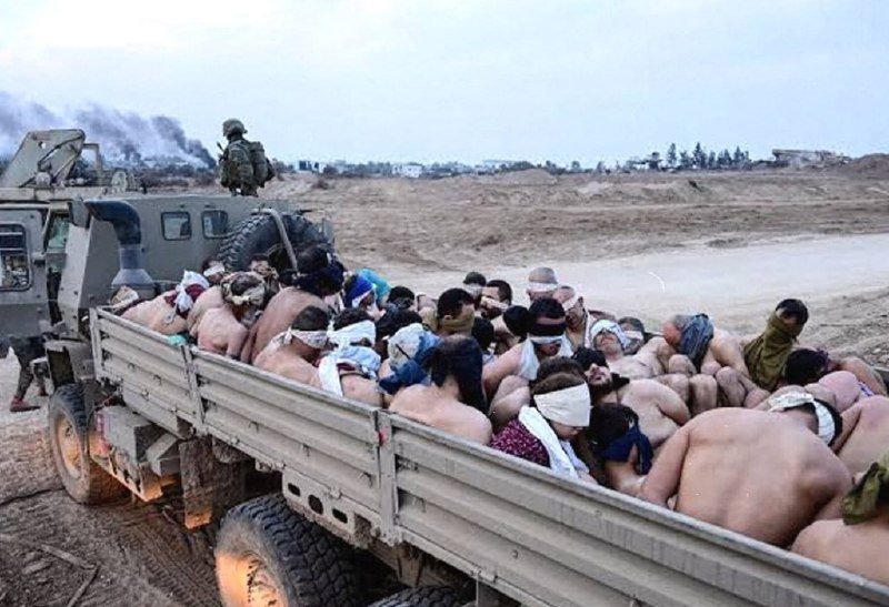 A military vehicle carries a large group of blindfolded, shirtless men through a barren landscape, with a soldier standing atop the vehicle.
