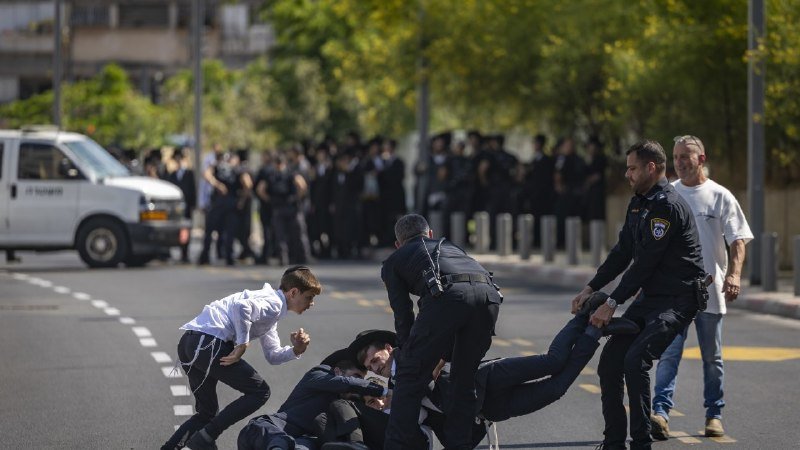 Uniformed officers restrain a person on a paved street while a group of similarly dressed individuals stands in the background near a white van and trees.