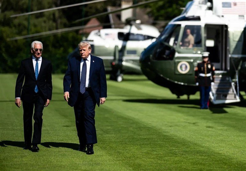 Two men in dark suits walk across a green lawn, with two helicopters—bearing U.S. flags and presidential seals—and a uniformed Marine visible behind them.