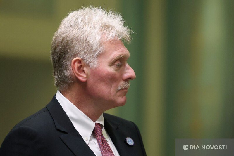 A white-haired man in a dark suit, white shirt, and red tie is shown in profile against a green background.