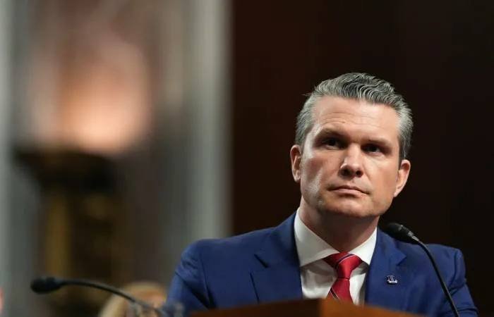 A man in a dark suit and red tie sits before microphones with a serious expression in a dimly lit room.