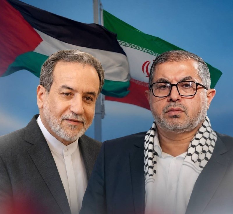 Two men pose together, with one wearing a Palestinian keffiyeh and the other in a dark suit, in front of the Palestinian and Iranian flags.