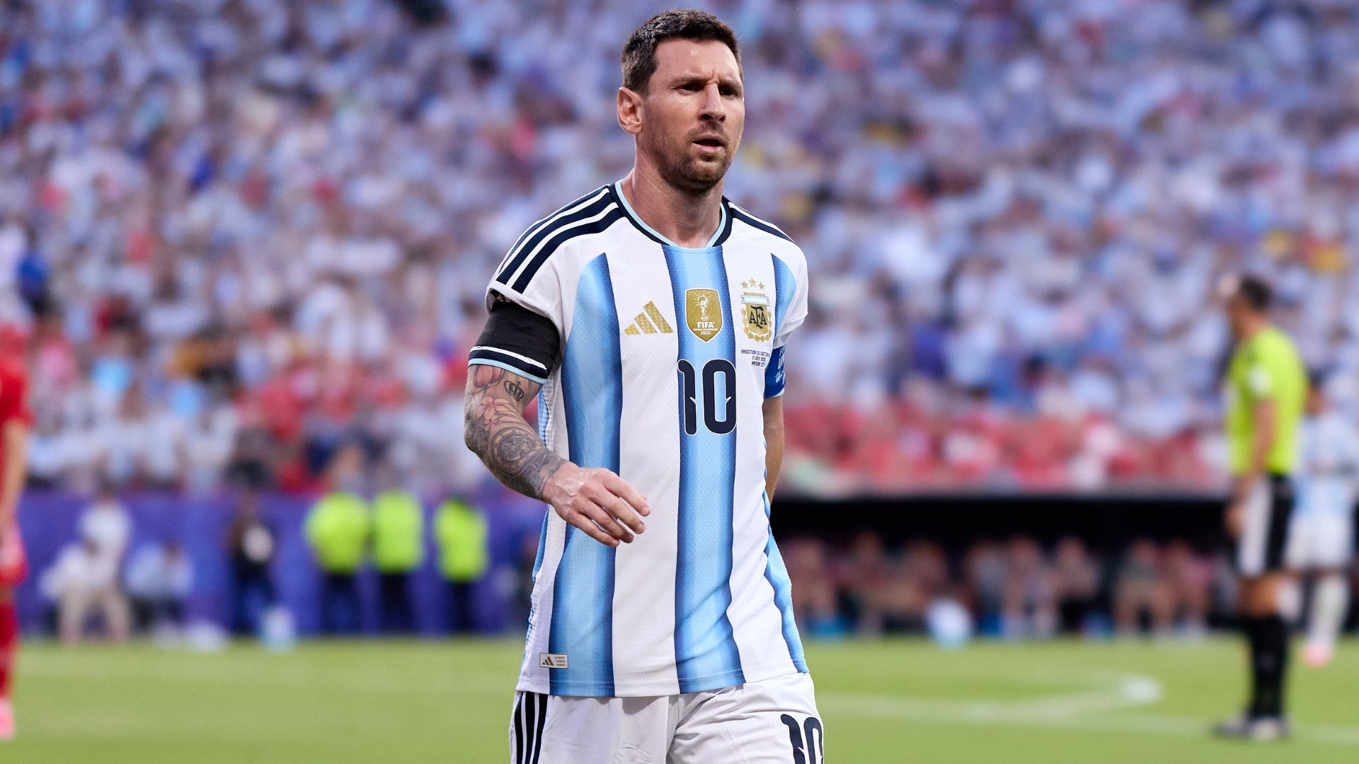 A bearded soccer player wearing a blue and white striped Argentina jersey with the number 10 stands on a stadium field before a blurred crowd.