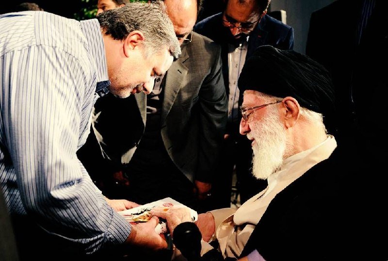 A younger man in a striped shirt smiles while handing a card or envelope to an elderly, white-bearded cleric wearing a black turban, as others observe.