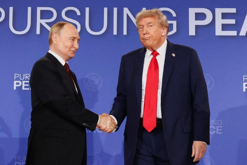 Two men in dark suits shake hands in front of a blue backdrop displaying the partially visible text "PURSUING PEA."