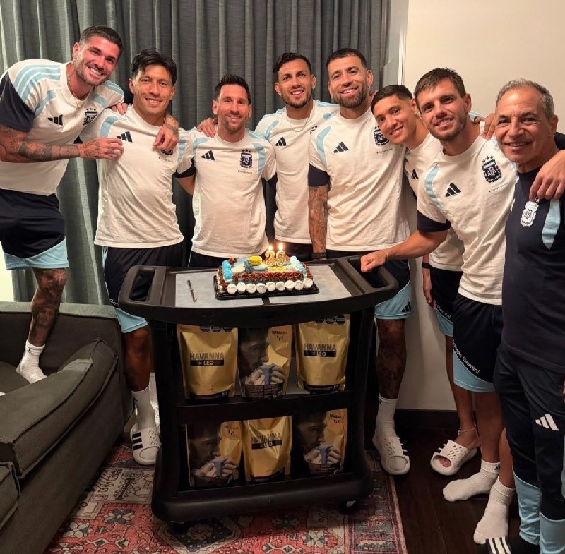 Eight men in white and light blue Argentina soccer jerseys pose smiling behind a cart displaying a lit birthday cake and "Havanna Leo" gift bags.
