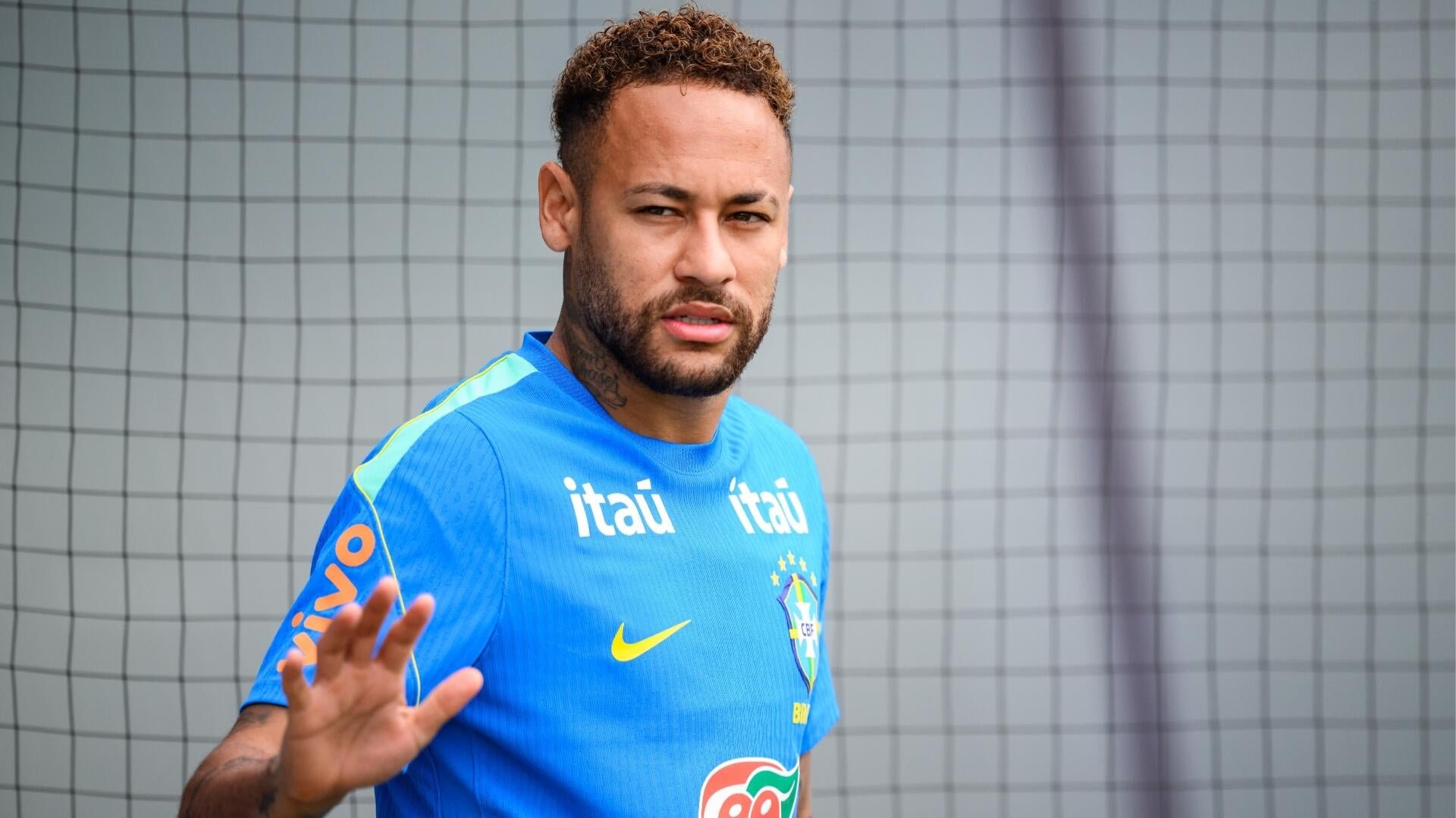 A man in a blue Brazil national team training jersey with "Itaú" sponsor logos raises his hand in front of a goal net.
