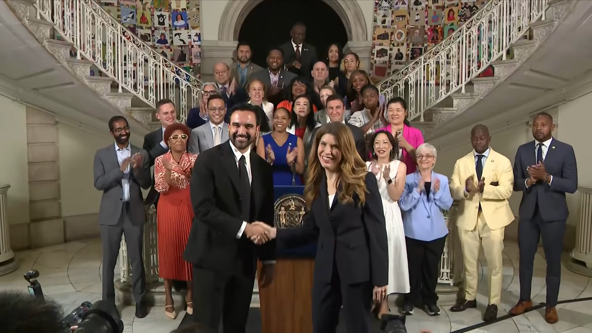 Two people shake hands at a wooden podium as a diverse group on a grand staircase applauds behind them in an ornate building.