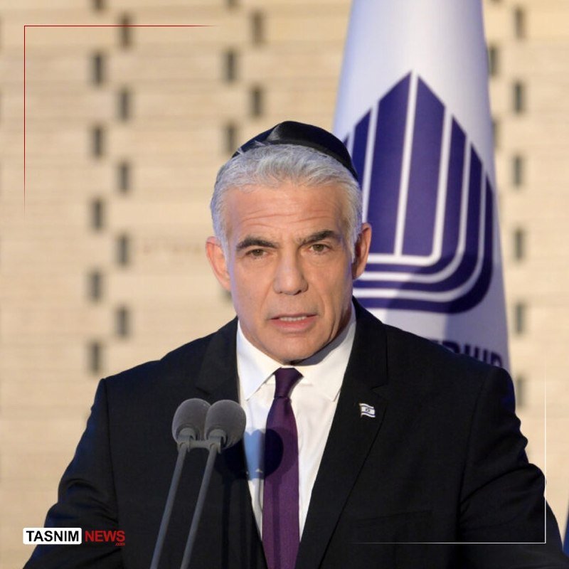 A gray-haired man wearing a black yarmulke, dark suit, and purple tie stands at a podium with two microphones, a flag visible behind him.