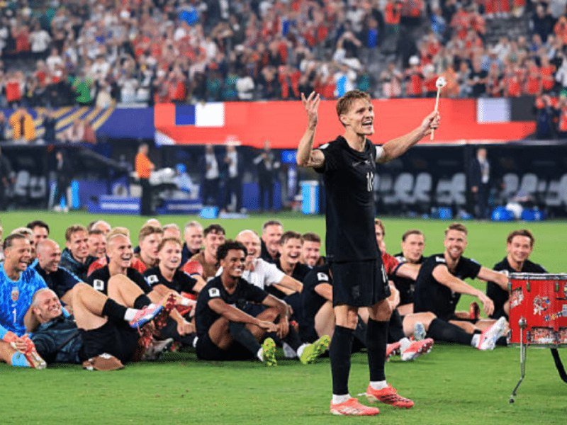 A soccer player in a black jersey celebrates with raised arms while teammates seated on the field and a cheering crowd cheer in a stadium.