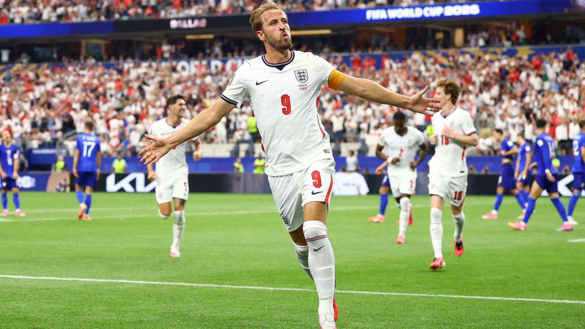 England's #9, wearing the captain's armband, celebrates with arms outstretched on the pitch during a soccer match.