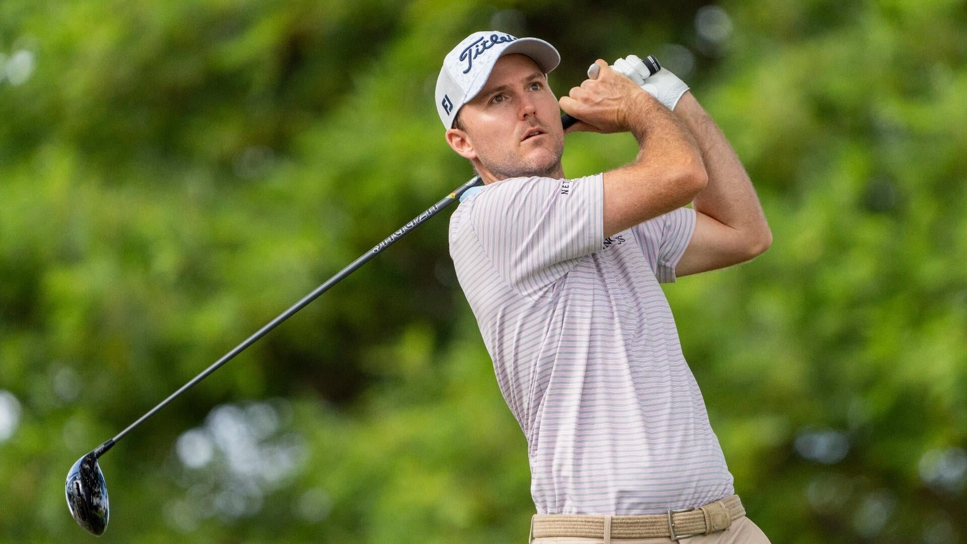 A golfer in a striped shirt and white Titleist cap follows through on a swing against a backdrop of green foliage.