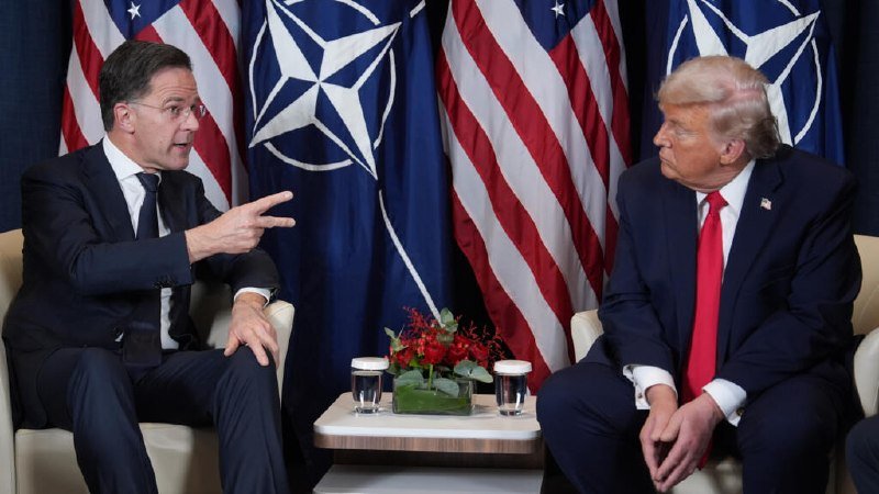 Two men in suits sit facing each other in armchairs, one gesturing while speaking, with U.S. and NATO flags displayed behind them.