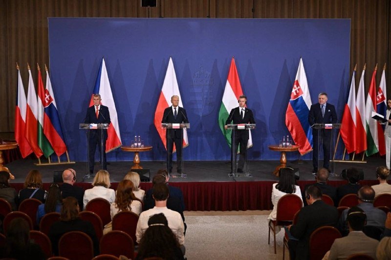 Four men in suits stand at separate podiums on a stage lined with Polish, Hungarian, Czech, and Slovak flags, addressing a seated audience in a formal conference setting.