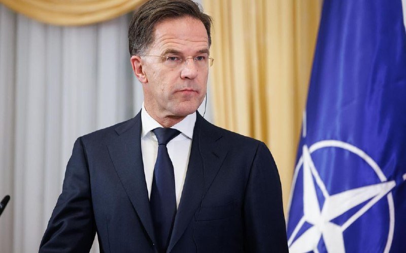 A man in a dark suit and tie stands beside a blue flag bearing a white compass-like emblem.