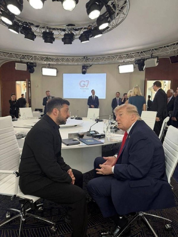 Two men in suits sit facing each other at a conference table beneath a screen displaying "G7 FRANCE," surrounded by overhead stage lights and other attendees.