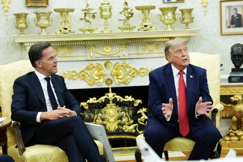 Two men in dark suits sit in ornate yellow armchairs in a gold-decorated room, with one gesturing while speaking during what appears to be a meeting.