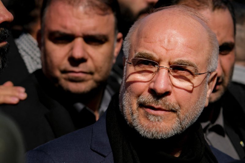 A gray-haired, bearded man wearing glasses and a dark coat stands surrounded by other men in suits under bright sunlight.