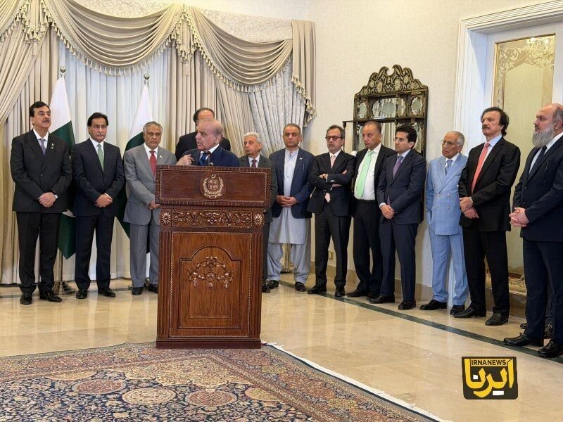 A man speaks at an ornate wooden podium bearing an emblem, flanked by about a dozen men in suits and traditional attire, with a flag visible behind them.