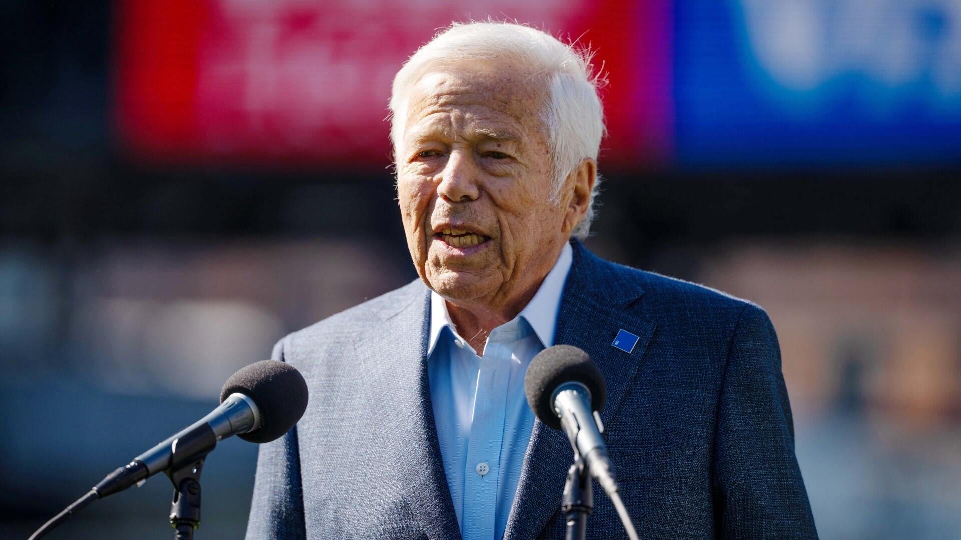 An elderly man with white hair, wearing a dark blazer and light blue shirt, speaks into two microphones at an outdoor podium with a blurred red and blue backdrop.