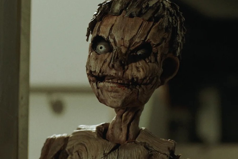 A figure with a grotesque, wood-like textured face, wide milky eyes, and exposed teeth stands in a dimly lit interior space.