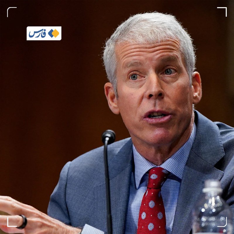 A gray-haired man in a blue suit and red tie speaks into a microphone, gesturing with his hand, with a water bottle visible nearby.