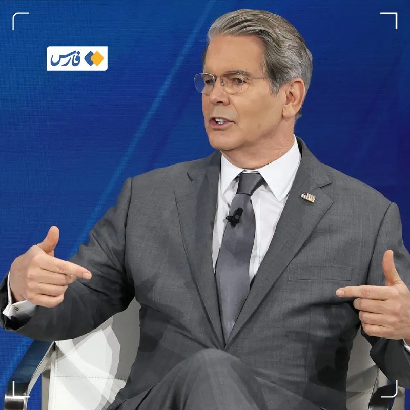 A gray-haired man in a gray suit, white shirt, and dark tie gestures with both hands while speaking during what appears to be a televised interview against a blue backdrop.