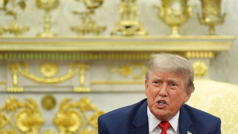 A man in a navy suit and red tie speaks while seated in an ornately decorated room with gold ornamental fixtures in the background.