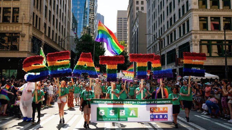 Parade participants in green shirts carry rainbow-floral letters spelling "SEATTLE" and a GSBA banner down a city street lined with crowds and tall buildings.