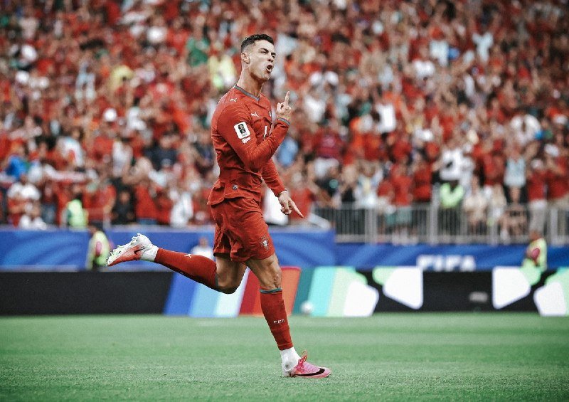 A soccer player in a red uniform runs on the field with his finger raised in celebration, as a large crowd in red cheers behind him.