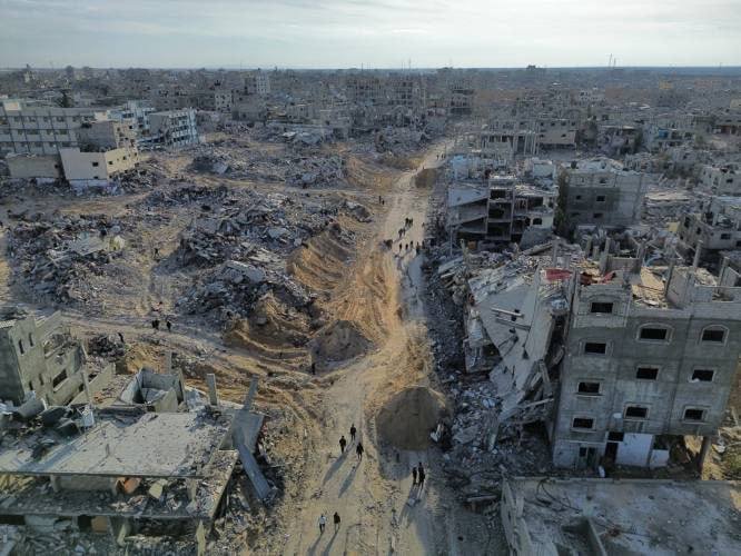 An aerial view shows a heavily destroyed urban area with collapsed buildings, widespread rubble, large craters, and small figures of people walking along a dirt path through the devastation.