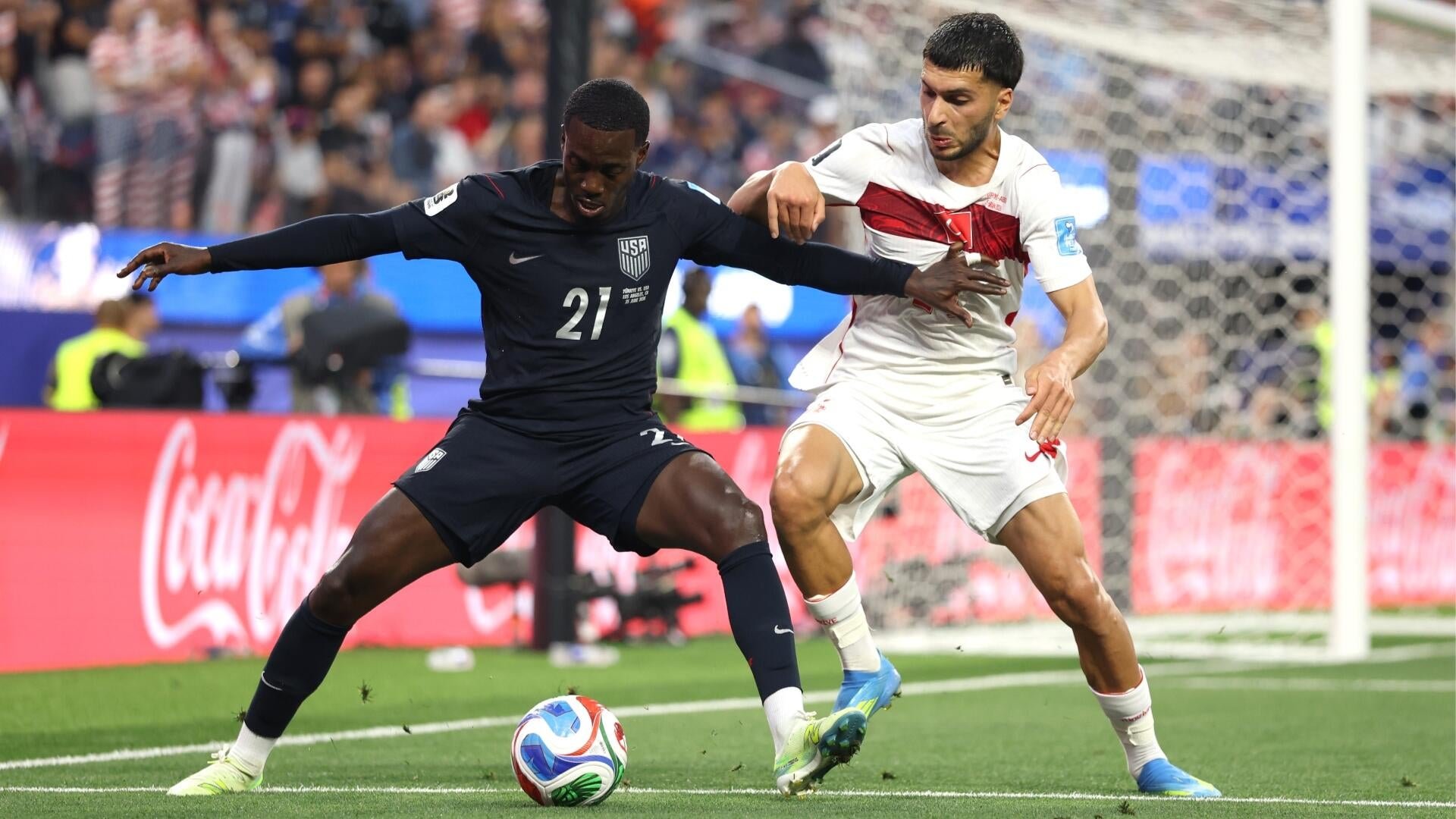 Two soccer players in dark and white jerseys compete for the ball near the goal net during a match in a stadium.