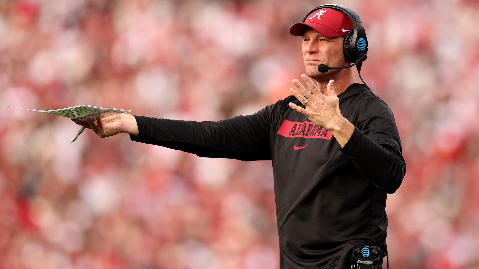 A coach in a black Alabama shirt and crimson cap wears a headset while gesturing with one hand and holding a play sheet and pen in the other, set against a blurred crowd.