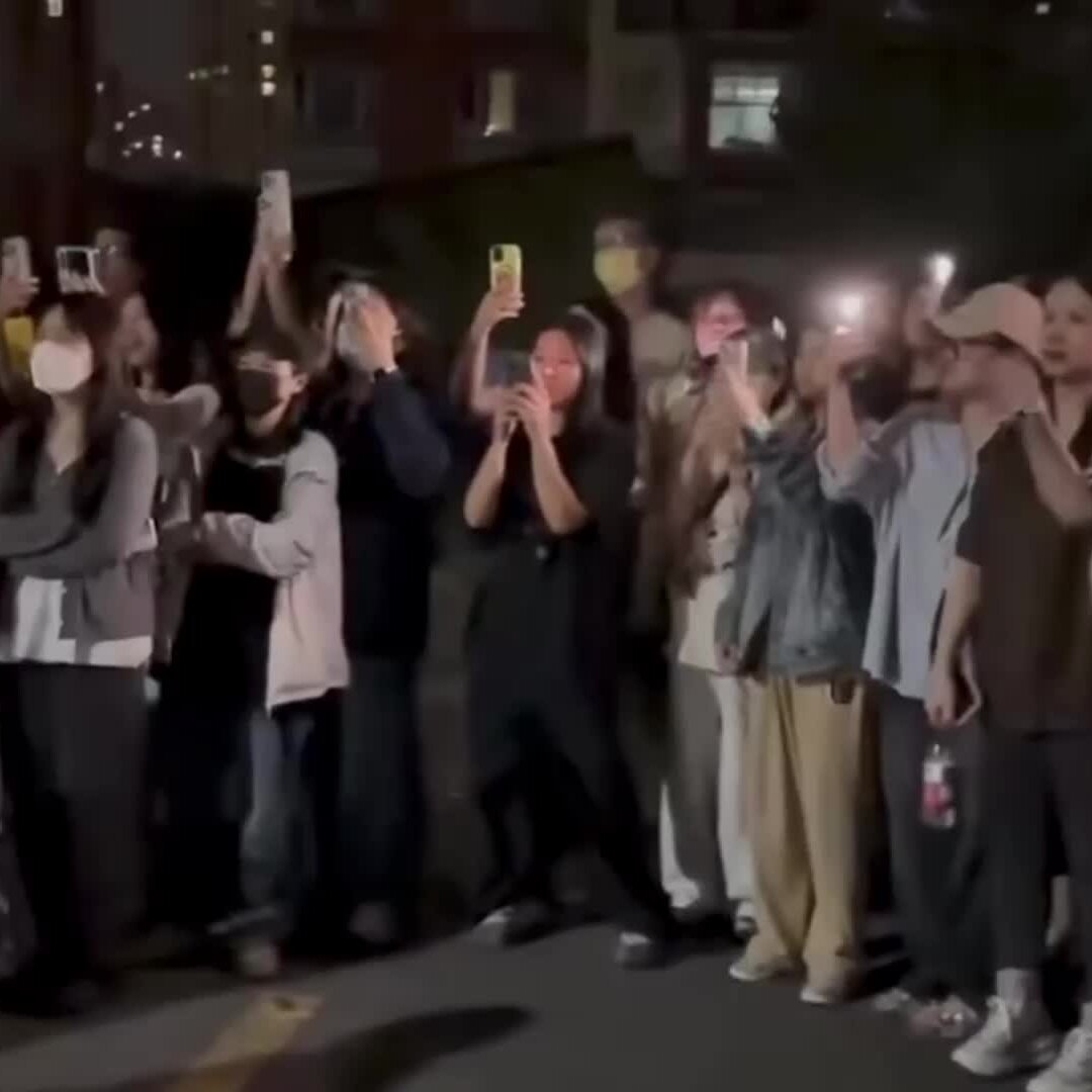 A nighttime street crowd holds up smartphones with flashlights on, recording something out of frame.