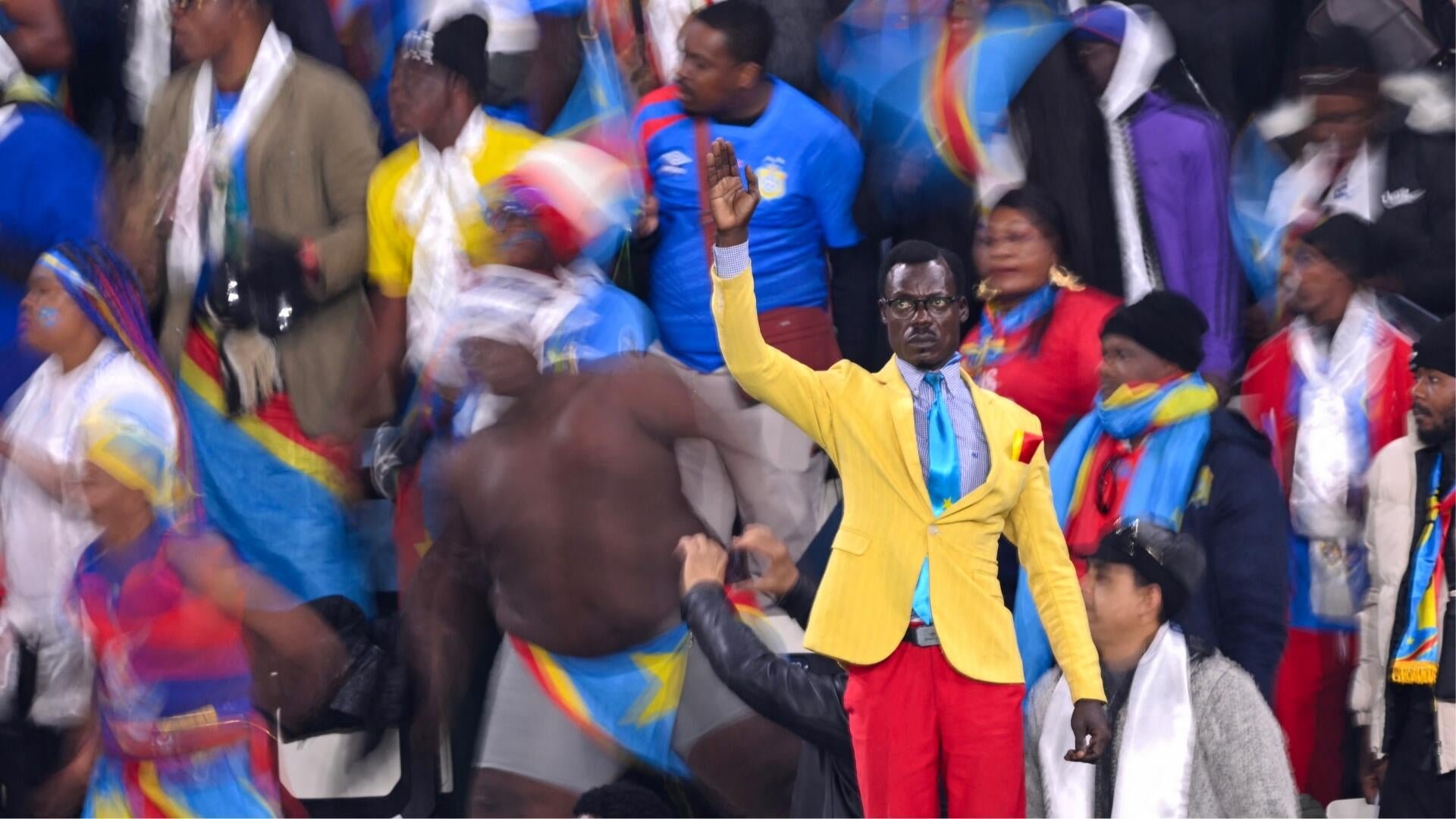 A man in a yellow blazer waves to a crowd, surrounded by people wearing colorful clothing and Congolese flags at an outdoor gathering.