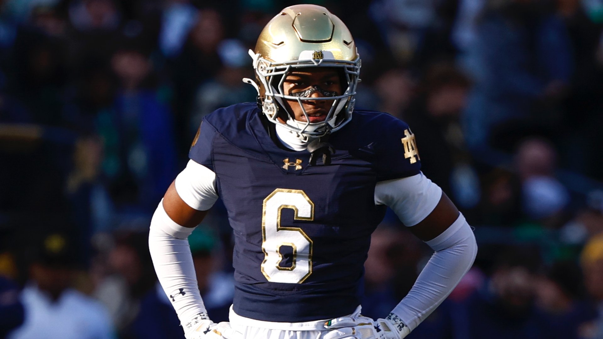 A football player wearing a navy blue jersey with the number 6 and a gold helmet stands on the field during a game, with a blurred crowd visible in the background.