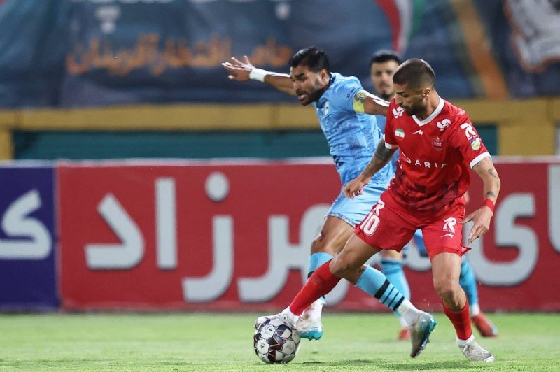 Two soccer players, one in light blue and one in red, compete for the ball on a grass field with advertising boards in the background.